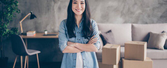 A young Asian woman stands with her arms crossed, smiling in front of a work desk, a couch, and sealed cardboard boxes.