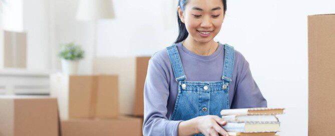 A young Asian woman is surrounded by other packed boxes as she packs a stack of books into a cardboard box