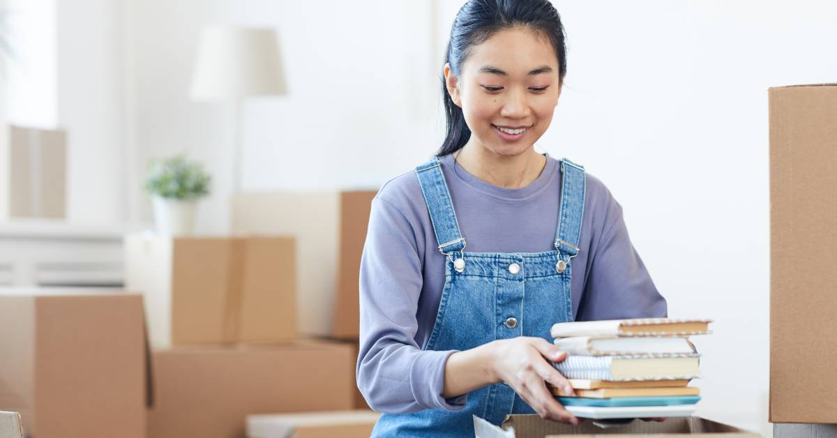 A young Asian woman is surrounded by other packed boxes as she packs a stack of books into a cardboard box
