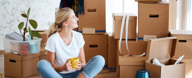 A young woman sitting in her new apartment surrounded by packed cardboard boxes, holding a coffee mug and smiling.