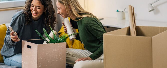 Two young women sit on a twin bed in an unpacked dorm room with two open cardboard boxes next to them looking at a phone.