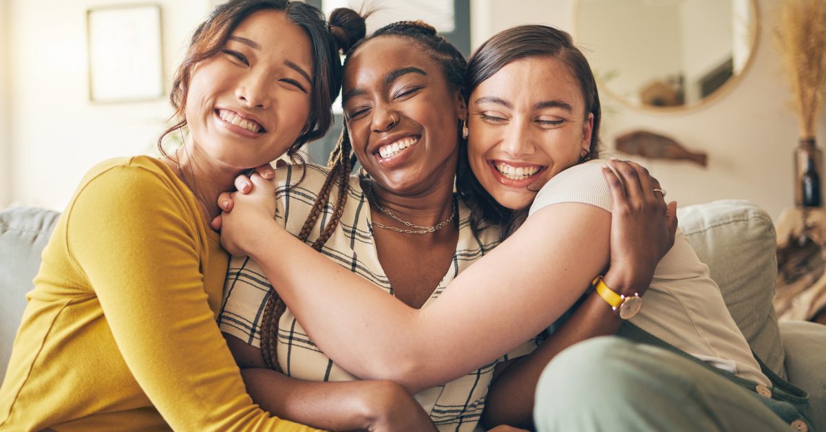 Three young women sit on a couch in a living room and wrap their arms around each other while smiling.