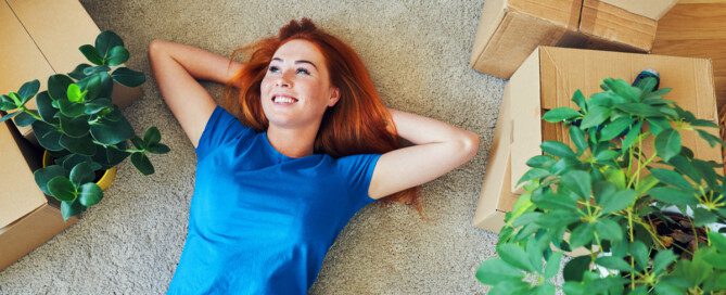 A young woman relaxes, lying on her carpet surrounded by a couch, house plants, and several cardboard boxes.