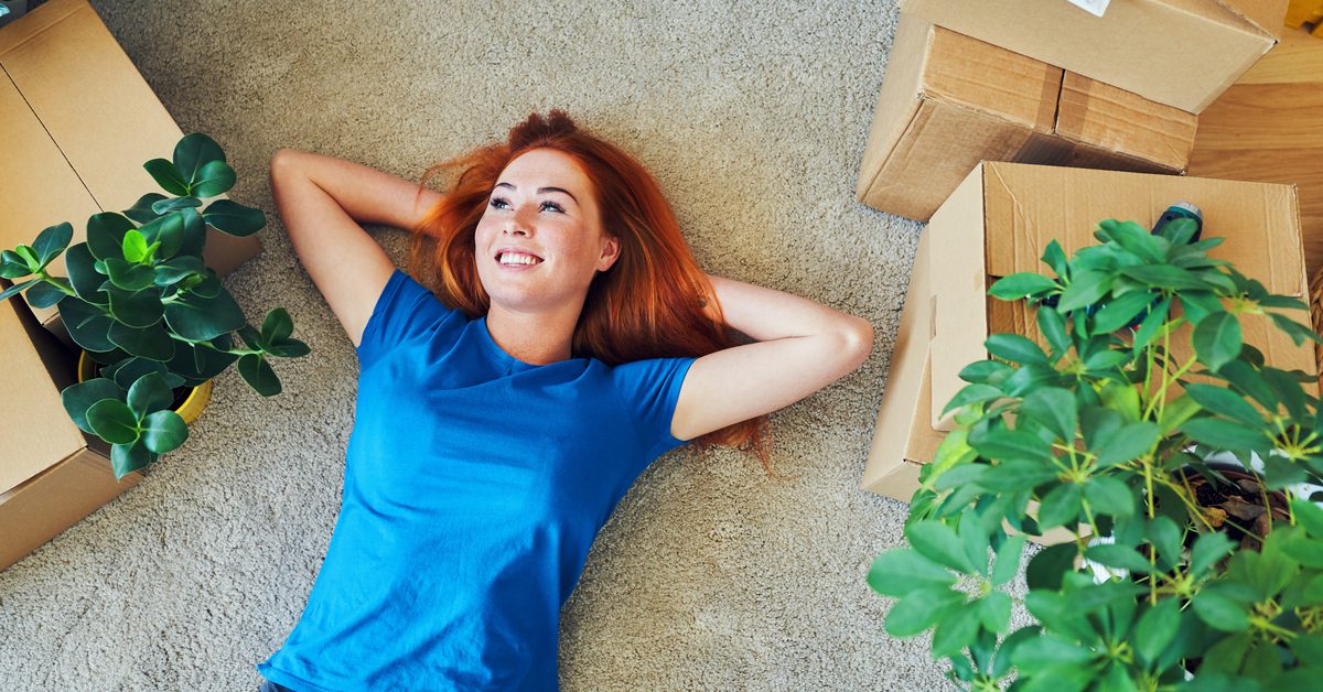 A young woman relaxes, lying on her carpet surrounded by a couch, house plants, and several cardboard boxes.