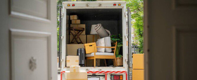 A moving truck filled with many cardboard boxes, a chair, a plant, and a rug with cardboard boxes to the side.