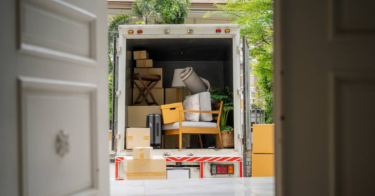 A moving truck filled with many cardboard boxes, a chair, a plant, and a rug with cardboard boxes to the side.