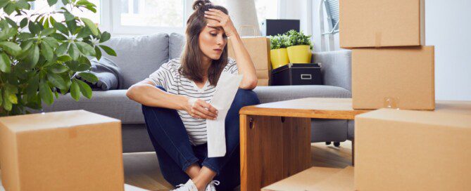 A woman sits on her apartment floor with her head in her hand, looking at a receipt and appearing stressed.