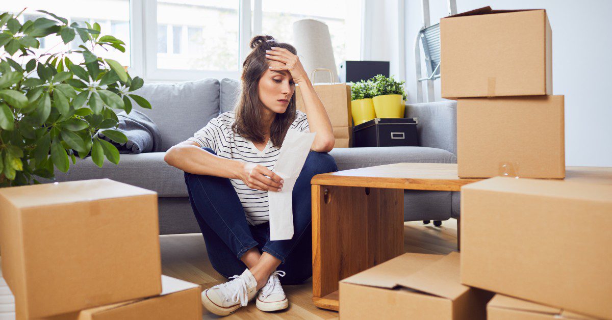 A woman sits on her apartment floor with her head in her hand, looking at a receipt and appearing stressed.