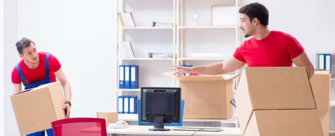 Two professional movers wearing red shirts transporting cardboard boxes out of a bright, furnished office.
