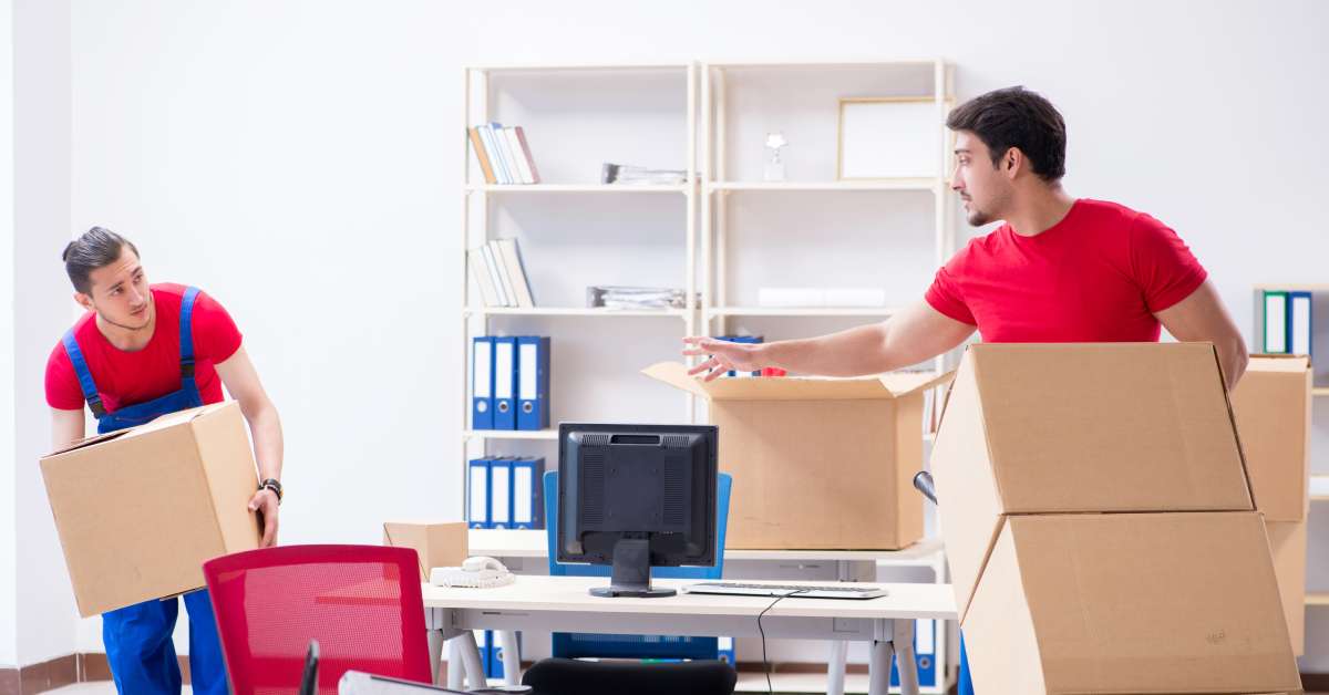 Two professional movers wearing red shirts transporting cardboard boxes out of a bright, furnished office.