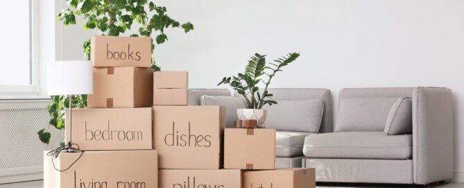 A stacked pile of packed and labeled cardboard boxes of various sizes with a grey couch in the background.