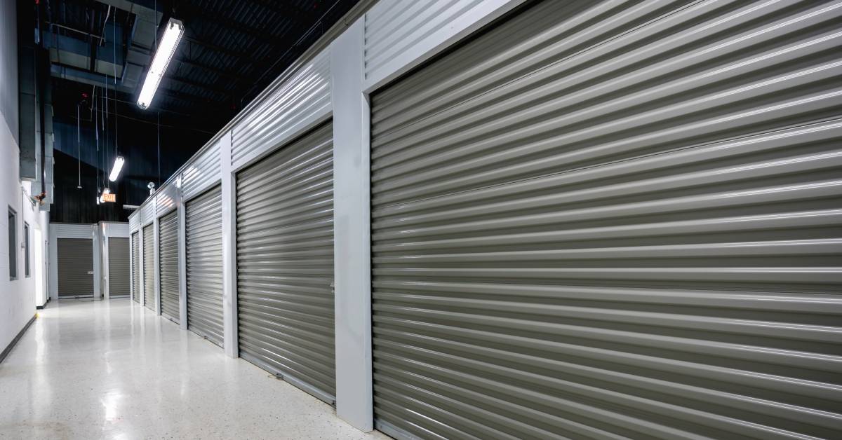 The hallway of an indoor storage facility with grey metal doors, light-colored tile flooring, and overhead fluorescent lights.