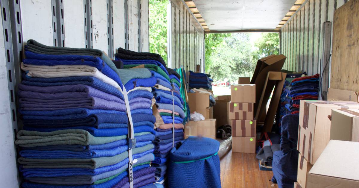 A moving trailer with objects wrapped in moving tarps and secured with straps near stacks of cardboard boxes.